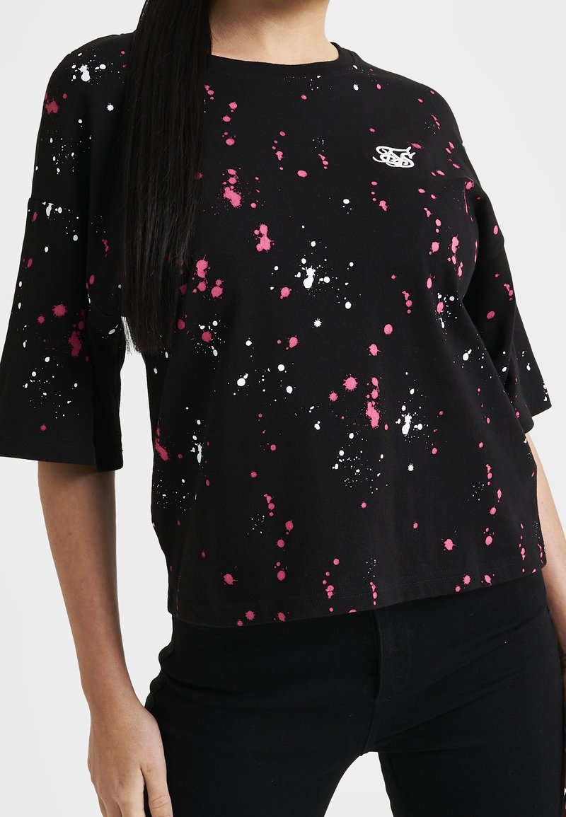 Mujer con una camiseta negra de manga corta con patrones de salpicaduras de pintura en rosa y blanco, y un pequeño logotipo blanco en el pecho.