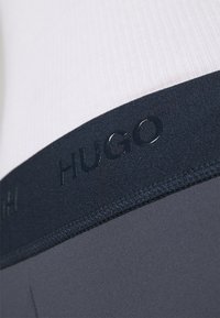 Tmavě modrý elastický pás s logem "HUGO", spárovaný s hladkým šedým materiálem. Detailní záběr zaměřující se na texturu a designové detaily.