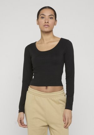 Žena s tmavými vlasmi v čiernom crop tope s dlhým rukávom a béžových nohaviciach s vysokým pásom, stojaca pred svetlým jednoliatym pozadím.