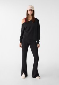 Schwarzer Off-Shoulder-Pullover mit langen Ärmeln, kombiniert mit schwarzen Locken-Hosen. Beigefarbenes Cap mit rotem Text und weißen Sneakers. Glatter Stofftextur.