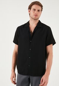 Camisa negra de manga corta con botón en la parte delantera, cuello clásico y corte relajado. La tela parece suave con una sutil textura.
