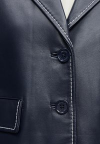 Gros plan sur une veste en cuir bleu marine avec des surpiqûres blanches contrastantes, deux boutons et une poche à rabat sur le côté gauche.