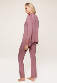 Dvodjelni loungewear komplet u nježnoj ružičastoj boji s čipkastim detaljima na rukavima i rubovima hlača. Ima opušteni kroj i duge rukave.