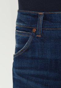Jeans de mezclilla azul oscuro con costuras naranjas prominentes, un bolsillo frontal y un detalle de botón de cobre en la cintura.