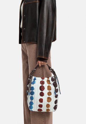 Osoba trzyma białą plisowaną torebkę z niebieskimi, czerwonymi, pomarańczowymi i fioletowymi okrągłymi wzorami oraz brązowymi plecionymi uchwytami.