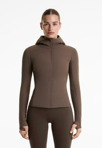 Jachetă maro, potrivită, cu glugă și fermoar complet. Fabricată dintr-un material neted și elastic. Prezintă orificii pentru degetul mare în mâneci pentru un plus de confort.