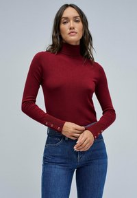 Pull à col roulé bordeaux avec des manches longues ajustées et des détails de boutons aux poignets, fabriqué à partir d'un tissu doux et extensible. Associé à un jean bleu.