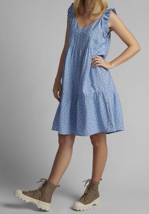 Kvinde iført en blå, ærmeløs kjole med hvide prikker og beige snørestøvler ved anklerne, stående foran en grå baggrund.