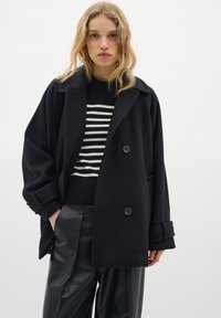 Black oversized wool coat with wide lapels and two buttons, layered over a black and white striped sweater, paired with black leather pants.