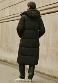 Musthave puffer-jakk mustaga kapuutsiga, millel on tikitud tekstuur ja pikk siluett. Kombineeritud mustade pükstega ja hõbedaste tossudega.