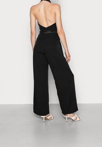 Jumpsuit noir avec un design décolleté dans le dos, style pantalon large, et accent satin à la taille. Modèle portant des talons nus à lanières.