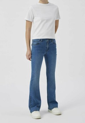 Person trägt ein einfaches weißes kurzärmliges T-Shirt, blaue ausgestellte Jeans und weiße Turnschuhe und steht vor einem hellgrauen Hintergrund.