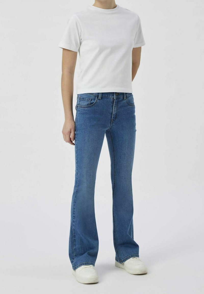 Personne portant un t-shirt blanc uni à manches courtes, un jean évasé bleu et des baskets blanches, debout devant un fond gris clair.