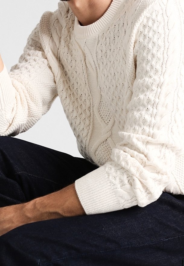 Homme assis portant un pull en maille torsadée crème texturé avec les manches retroussées et un jean bleu foncé sur un fond uni.
