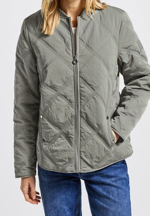 Vrouw die een lichtgrijs gewatteerd jack met diagonale stiksels en een ritssluiting draagt, gecombineerd met donkerblauwe spijkerbroek, één hand in de zak, tegen een effen achtergrond.