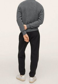 Grauer gestrickter Pullover mit gerippten Bündchen, kombiniert mit schwarzen Jogginghosen. Das Model trägt weiße Turnschuhe und hat die Hände hinter dem Rücken verschränkt.