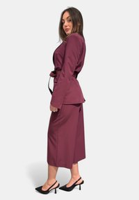 Completo su misura bordeaux con giacca aderente, pantaloni a gamba larga e una cintura nera. Abbinato a décolleté neri con slingback. Texture del tessuto liscia.