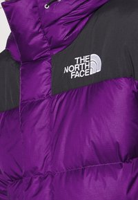 Paarse pufferjas met zwarte accenten, voorzien van een hoge kraag, doorgestikte textuur en het witte geborduurde logo van "The North Face".
