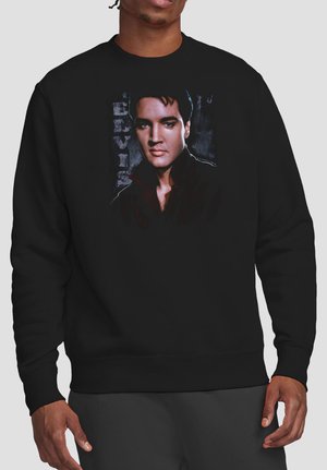 Mann trägt schwarzen Pullover mit farbigem Porträt eines Mannes und vertikalem Text "ELVIS" auf der Brust.
