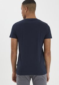 Tumši zila apģērba t-kreklā ar apļveida kaklu, kas izgatavots no kokvilnas, īsām piedurknēm, gludu faktūru un taisnu apakšmalu. Aizmugures skats, minimāla apdare un bez logotipiem.