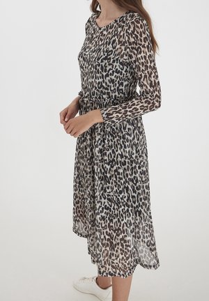 Mujer con vestido de manga larga hasta la rodilla con estampado de leopardo y tela transparente, combinado con zapatillas blancas, de pie frente a un fondo liso.