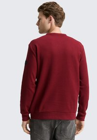 Felpa rossa girocollo in tessuto testurizzato, con maniche lunghe e polsini a costine. Presenta un colore uniforme con una toppa del logo sulla manica.