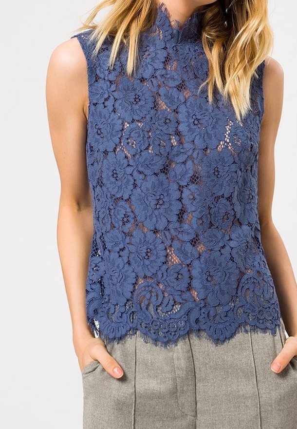 Top de encaje azul marino con patrón floral y dobladillo en forma de ondas. Diseño sin mangas con cuello alto y textura transparente. Combinado con pantalones grises.