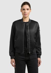 Femme debout portant une veste bomber en satin noir avec fermeture éclair à l'avant et un pantalon noir, sur un fond gris clair.