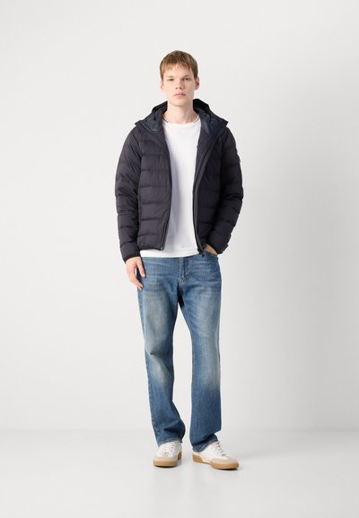 Doudoune navy avec une capuche, présentant une texture matelassée. Associée à un jean bleu et des baskets blanches avec des semelles beige. T-shirt blanc en dessous.