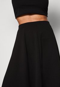 Černá áčková sukně z texturované látky, s úzkým pasem a sklady, která ukazuje jednobarevný design bez dalších vzorů.