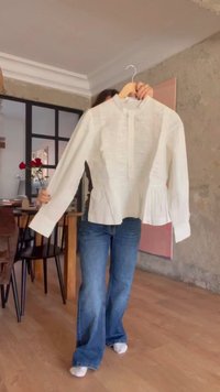 Osoba držící na ramínku bílou blůzu s dlouhým rukávem v jídelně s dřevěnou podlahou a nábytkem.