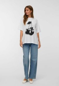 Witte oversized t-shirt met een zwart kersenafbeelding, gecombineerd met lichtblauwe wijde jeans en beige platte schoenen. Eenvoudig, casual ontwerp.