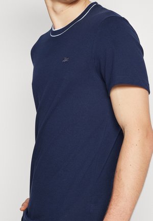 Homme portant un t-shirt bleu marine à manches courtes avec un liseré blanc au col et un petit logo brodé sur la poitrine.