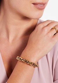Goldene Kettenarmband mit ineinander greifenden Gliedern und glänzender Oberfläche, am Handgelenk getragen, kombiniert mit einem goldenen Ring, vor einem sanften rosa Oberteil.