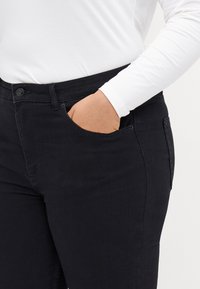 Svarte denimjeans med en tettsittende design, med en frontlomme og sydd søm. Brukt sammen med en hvit langermet skjorte.