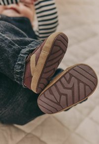 Des chaussures à semelle souple présentant un design marron et beige, avec une semelle en caoutchouc texturé et un motif cousu près du talon. Un jean en denim est visible.