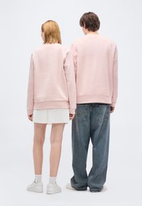 Δύο άτομα στέκονται με την πλάτη στην κάμερα, φορώντας αντίστοιχα ανοιχτοροζ φούτερ. Το ένα φορά λευκή πλισέ φούστα και αθλητικά παπούτσια, ενώ το άλλο φαρδιά τζιν.