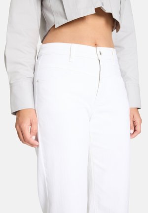 Frau trägt weiße Jeans mit hoher Taille und ein kurzes hellgraues Shirt, das den Bauch zeigt, und die Hände ruhen seitlich.