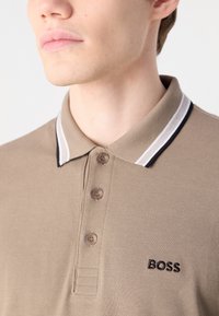 Pólo bege com um tecido texturizado, três botões castanhos e um colarinho riscado em branco e preto. Apresenta o logótipo "BOSS" em preto.