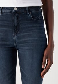 Vysoké tmavě modré džíny z denimu s jemnou texturou, se zadními kapsami a stříbrným zapínáním na knoflík.
