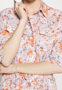 Camisa floral que presenta una mezcla de colores naranja, lavanda y aguamarina, con mangas cortas, bolsillos con botones y una cintura ajustada con pasadores para cinturón.