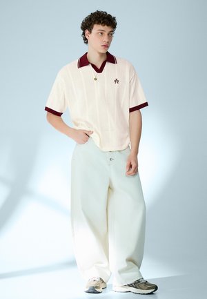 Persona giovane con capelli ricci che indossa pantaloni ampi color crema, maglia di maglia color crema con colletto e polsini bordeaux, e sneakers beige, in piedi con la mano in tasca.