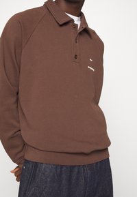 Polo a maniche lunghe marrone con colletto a costine, chiusura a due bottoni e logo ricamato. Il materiale sembra morbido con una finitura testurizzata.