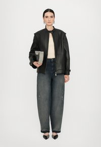 Jachetă din piele neagră cu guler înalt, bluză bej, blugi de denim albastri supradimensionați și pantofi cu toc ascuțit în nuanțe de negru și gri. Ține în mână o poșetă cu mărgele.