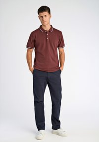 Bordowa koszulka polo z kołnierzykiem i trójprzyciskowym paskiem, z kontrastowymi białymi lamówkami, zestawiona z granatowymi spodniami i białymi trampkami.