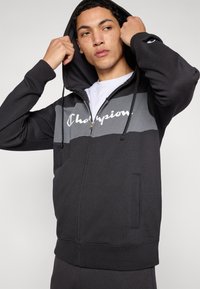 Μαύρο φούτερ με φερμουάρ και γκρι οριζόντιες ρίγες, με το λογότυπο "Champion". Διαθέτει κουκούλα με κορδόνι και πλαϊνές τσέπες, κατασκευασμένο από μαλακό ύφασμα.