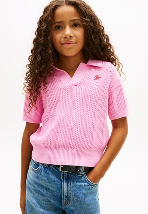 Ung person med krøllet hår iført en lyserød kortærmet striktrøje og blå jeans med et sort bælte, stående med den ene hånd i lommen.