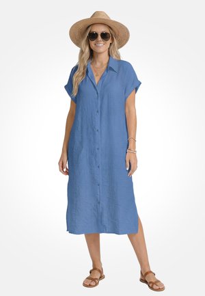 Kobieta w niebieskiej sukience midi zapinanej na guziki, nosząca słomkowy kapelusz, okulary przeciwsłoneczne, warstwowe naszyjniki, bransoletki i brązowe sandały, stojąca na białym tle.