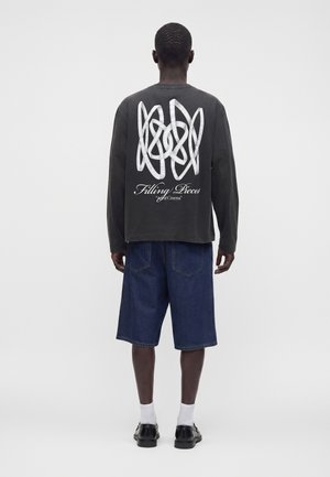 Mand iført sort langærmet skjorte med hvidt abstrakt mønster og teksten "Filling Pieces", blå denimshorts, hvide sokker og sorte sko, set bagfra.
