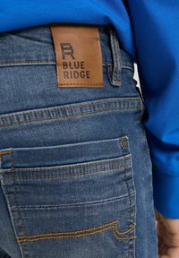 Modré džínové kalhoty s koženou nášivkou označenou "BLUE RIDGE". Disponují šitými kapsami a kontrastním oranžovým prošíváním.